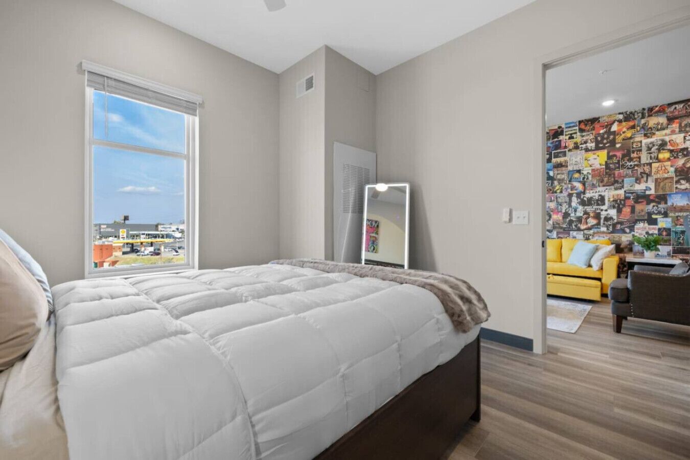 A bedroom with a large bed, a window with a city view, a mirror, and a wall covered with pictures.