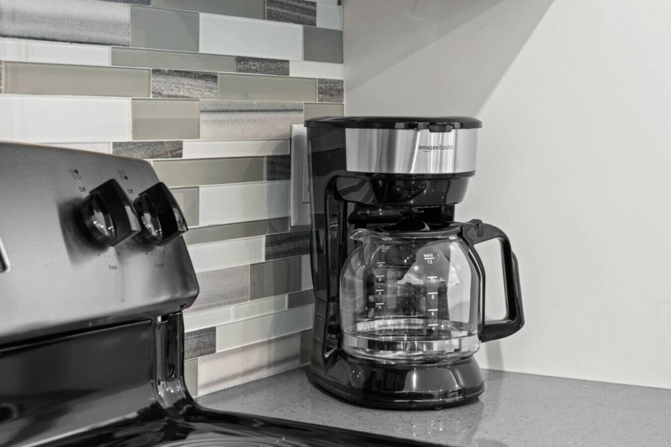 A coffee maker with a clear carafe and a black base, positioned on a countertop next to a tiled backsplash.
