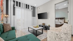 A modern living room with a green sofa, a coffee table, and a patterned armchair. A flat-screen TV is mounted on the wall, and a bed is visible through a glass door. - Music On in Nashville - 8 min from Broadway! (Nashville)