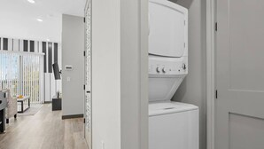 A modern laundry room with a front-loading washer and dryer, a wooden floor, and a view of the living area with a sofa and a table. - Music On in Nashville - 8 min from Broadway! (Nashville)