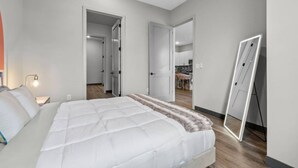 A bedroom with a bed, bedside tables, a mirror, and a door leading to a kitchen.