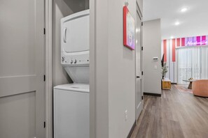 A laundry room with a washing machine and dryer, a hallway leading to a living area with a couch and a neon sign.