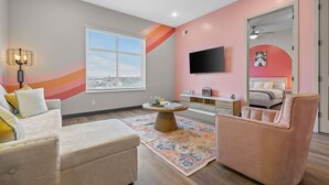 Un salon moderne avec un canapé, un fauteuil et une table basse. Un téléviseur est fixé au mur, il y a une fenêtre avec des stores et un tapis décoratif.
