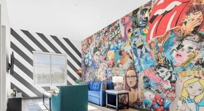 Una habitación con un gran mural de cómics en la pared, un sofá azul, una mesita con una lámpara y una ventana con persianas.