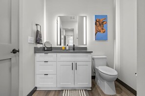 A modern bathroom with white cabinetry, a sink, and a toilet. There is a mirror above the sink and a painting of a cow on the wall. - Spacious 1BR for 4, Free WiFi | 8 Min to Broadway (Nashville)