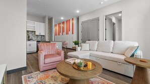 A modern living room with a white sofa, a pink armchair, and a round wooden coffee table. There is a kitchen in the background with white cabinets and a striped wall decoration. - Chic 2BR Getaway in Downtown Nashville! (Nashville)