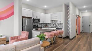 A modern kitchen with white cabinets, stainless steel appliances, and a gray backsplash. There are pink chairs and a round wooden table in the living area. - Chic 2BR Getaway in Downtown Nashville! (Nashville)