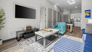 Una sala moderna con un televisor de pantalla plana, un sillón azul, una mesa de centro y un sofá. Se observa una cocina al fondo con armarios blancos y un lavamanos.