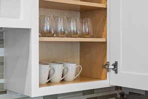 Un meuble de cuisine à deux étagères ; l’étagère supérieure contient des verres en verre transparent, et l’étagère inférieure est garnie de tasses blanches.