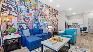 Un salon moderne agrémenté d’une fresque murale aux couleurs vives, d’un canapé bleu, d’une table basse et d’une cuisine en arrière-plan.