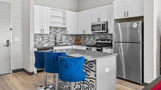 A modern kitchen with white cabinets, a stainless steel refrigerator, a microwave, and a stove. There are two blue bar stools at the island.