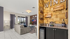 Un salón moderno con un sofá gris, una mesa de centro de vidrio y un televisor. Se ve una zona de cocina con una pared de madera y una estantería empotrada para vinos.