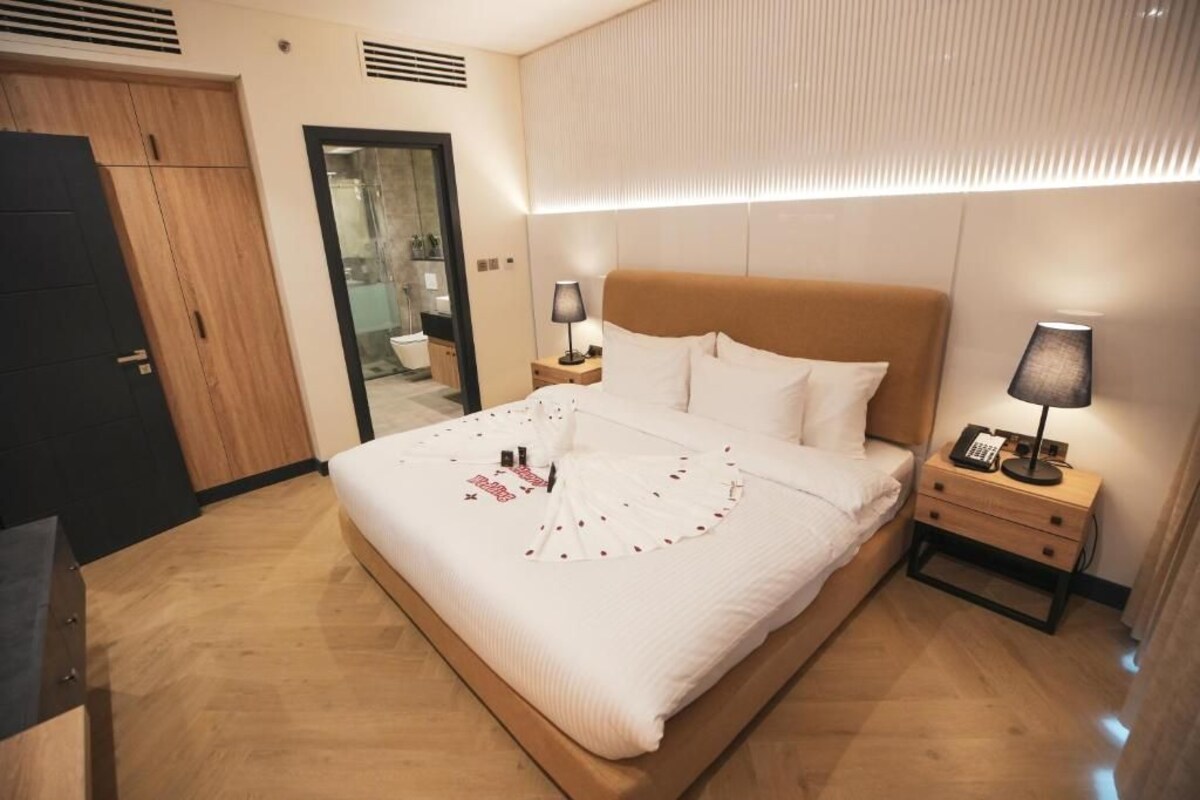 A neatly made bed with white linens and a decorative pillow arrangement. There is a bedside table with a lamp and a phone. The room has a wooden floor and a door to the left.