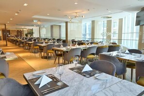 A spacious restaurant with marble-topped tables and modern chairs, large windows, and a high ceiling.