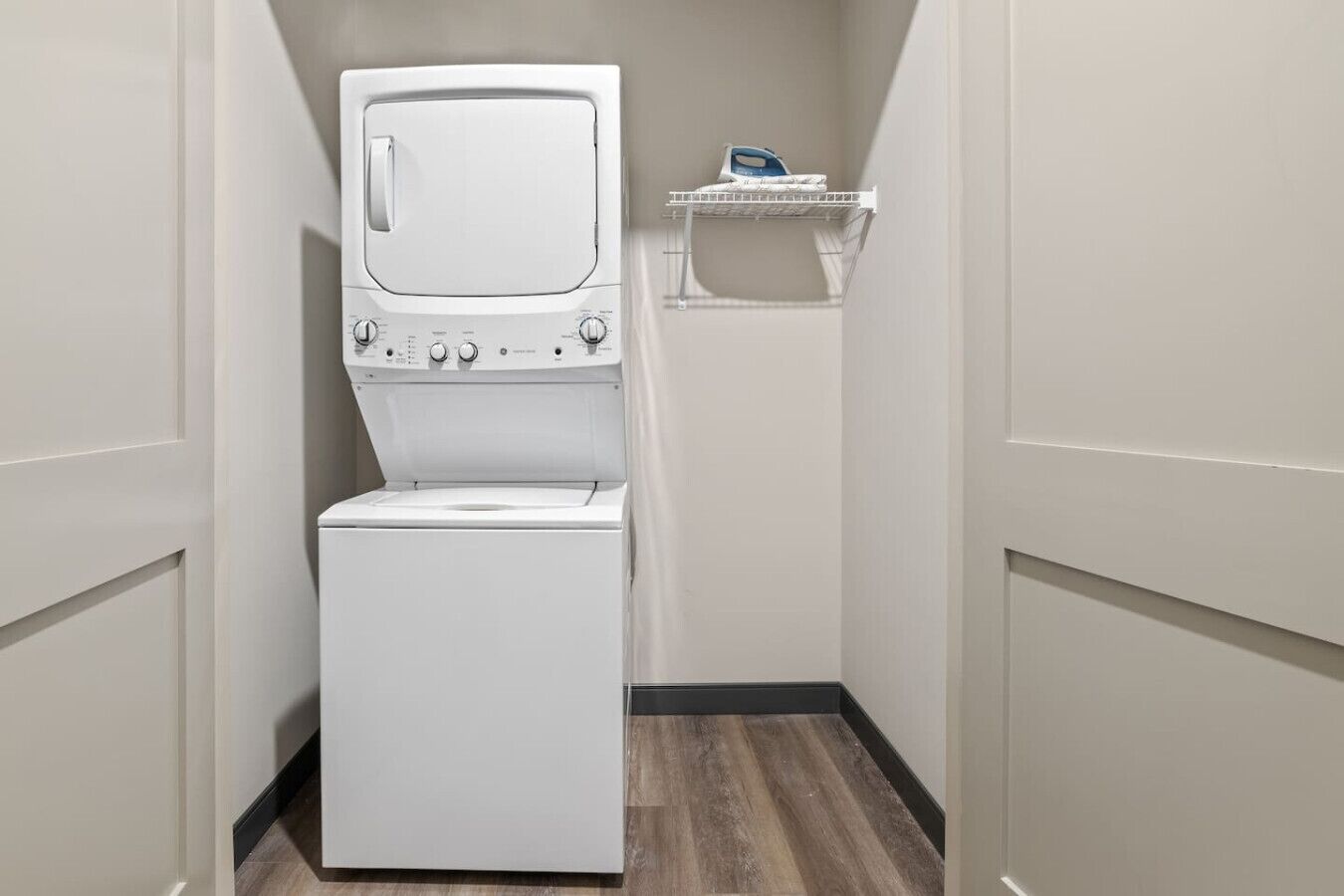 A compact laundry room with a front-loading washer and dryer, a small shelf with a blue item, and a wooden floor.