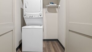 A compact laundry room with a front-loading washer and dryer, a small shelf with a blue item, and a wooden floor. - Perfect for Family Getaway - Mins from Music City! (Nashville)