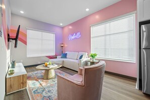 A modern living room with a pink accent wall, a neon sign, a white sofa, a pink armchair, and a wooden coffee table. - Perfect for Family Getaway - Mins from Music City! (Nashville)