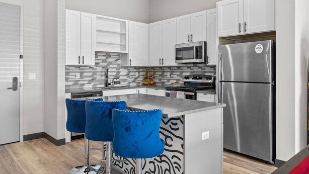 A modern kitchen with white cabinets, a stainless steel refrigerator, a microwave, and a gas stove. There are two blue bar stools at the island. - Hip 1BR Hideaway - Quick Drive to Downtown Fun! (Nashville)