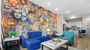 Un salon moderne agrémenté d’une fresque murale colorée de style pop art, d’un canapé bleu, d’un fauteuil turquoise et d’un petit coin cuisine visible au fond.