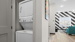 Une buanderie équipée d’un lave-linge et d’un sèche-linge à chargement frontal, avec un mur à rayures et un fauteuil turquoise dans l’espace de vie adjacent.