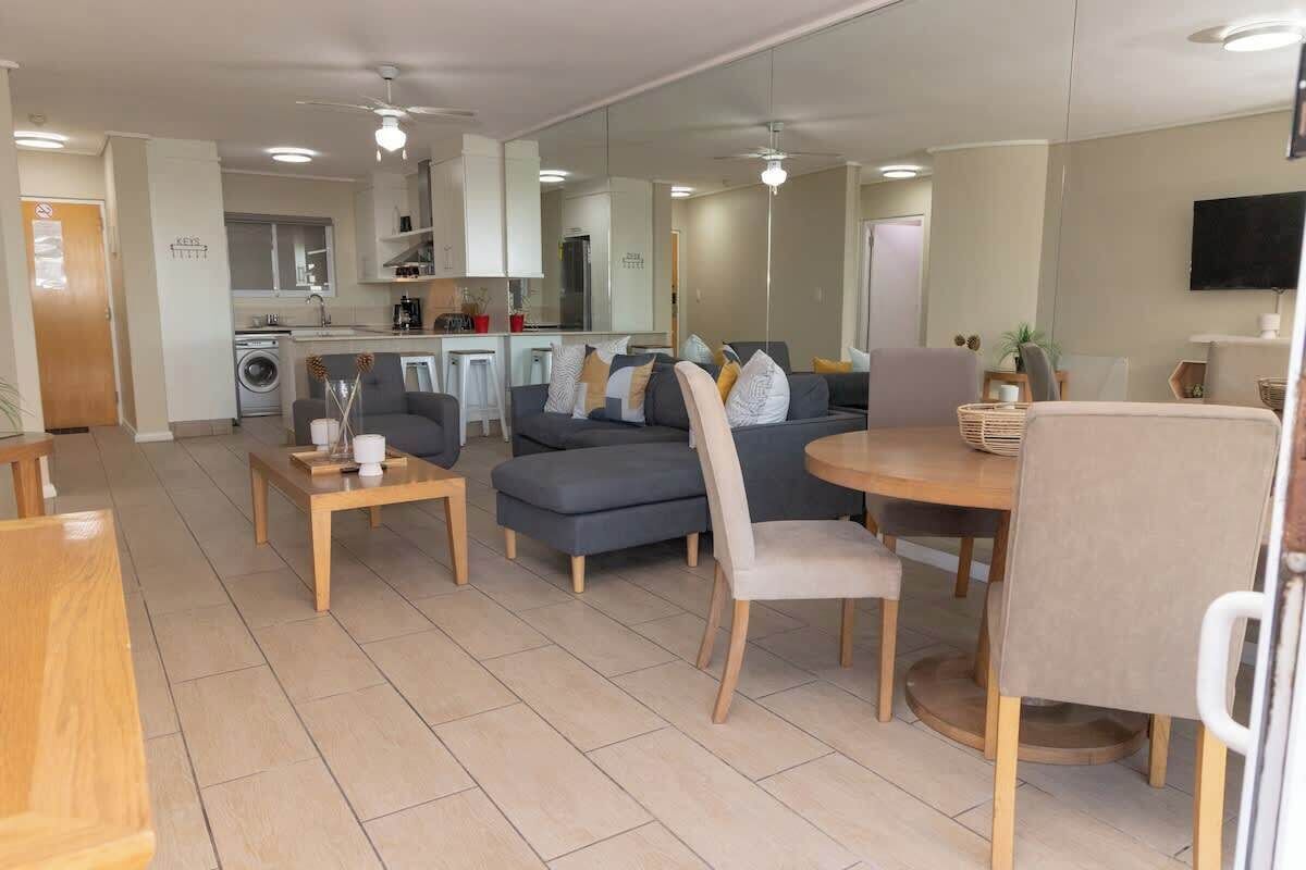 A modern living room with a sofa, chairs, a coffee table, and a dining table. There is a kitchen in the background with a washing machine and a microwave.
