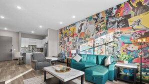 Un salon moderne agrémenté d’une fresque murale aux couleurs vives, d’un canapé turquoise et d’une cuisine au design minimaliste en arrière-plan.