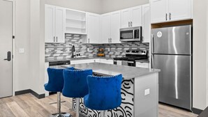 A modern kitchen with white cabinets, a stainless steel refrigerator, a microwave, and a gas stove. There are two blue bar stools at the island.