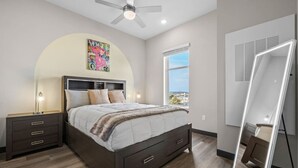 A modern bedroom with a bed, nightstands, a ceiling fan, a large window, and a mirror.
