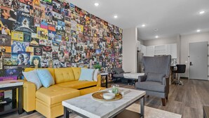 A living room with a yellow sofa, a grey armchair, and a coffee table with a tray. A wall covered in colorful posters and a small plant on the coffee table.