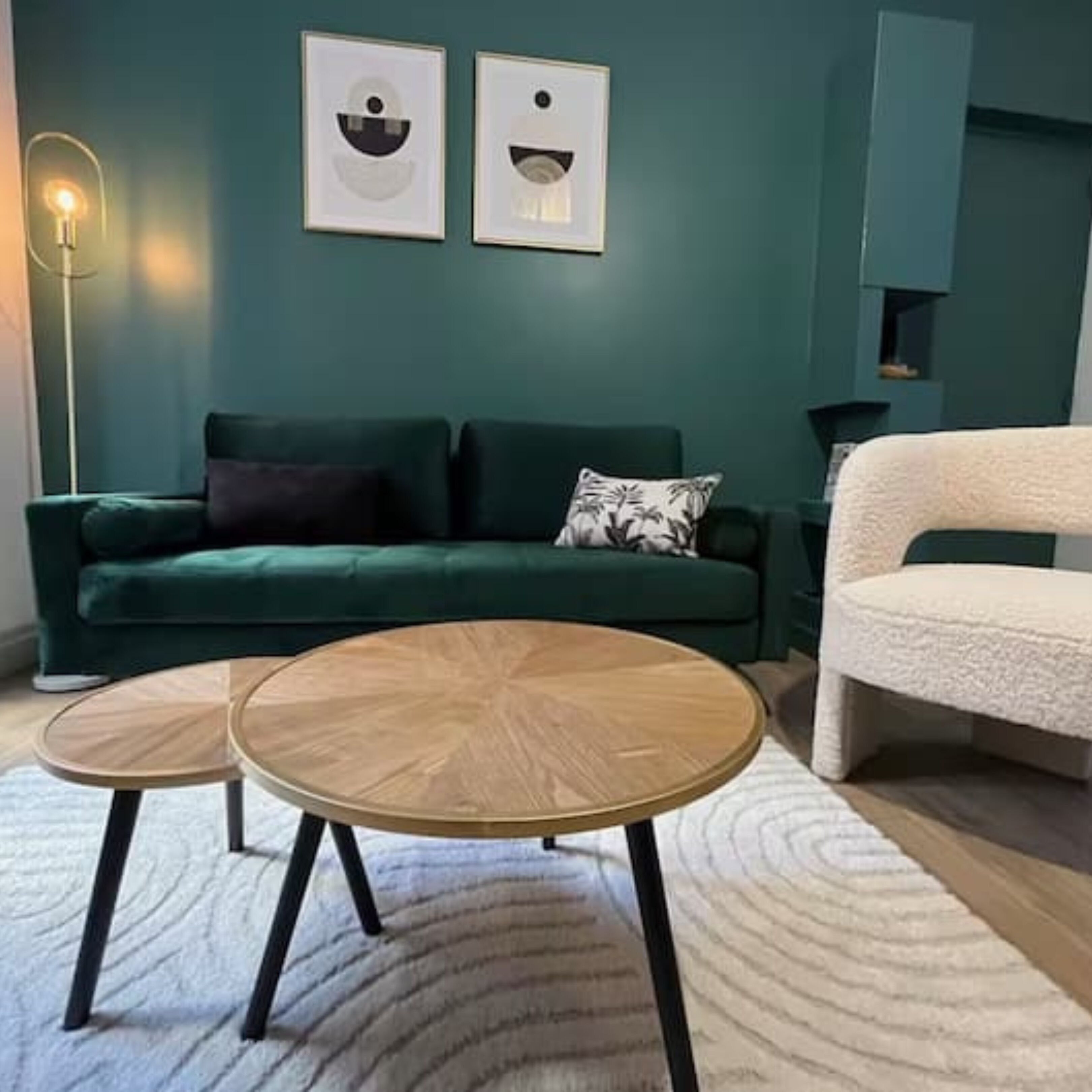 Una sala de estar con un sofá verde, dos mesas redondas de madera, un sillón blanco y una lámpara de pie.