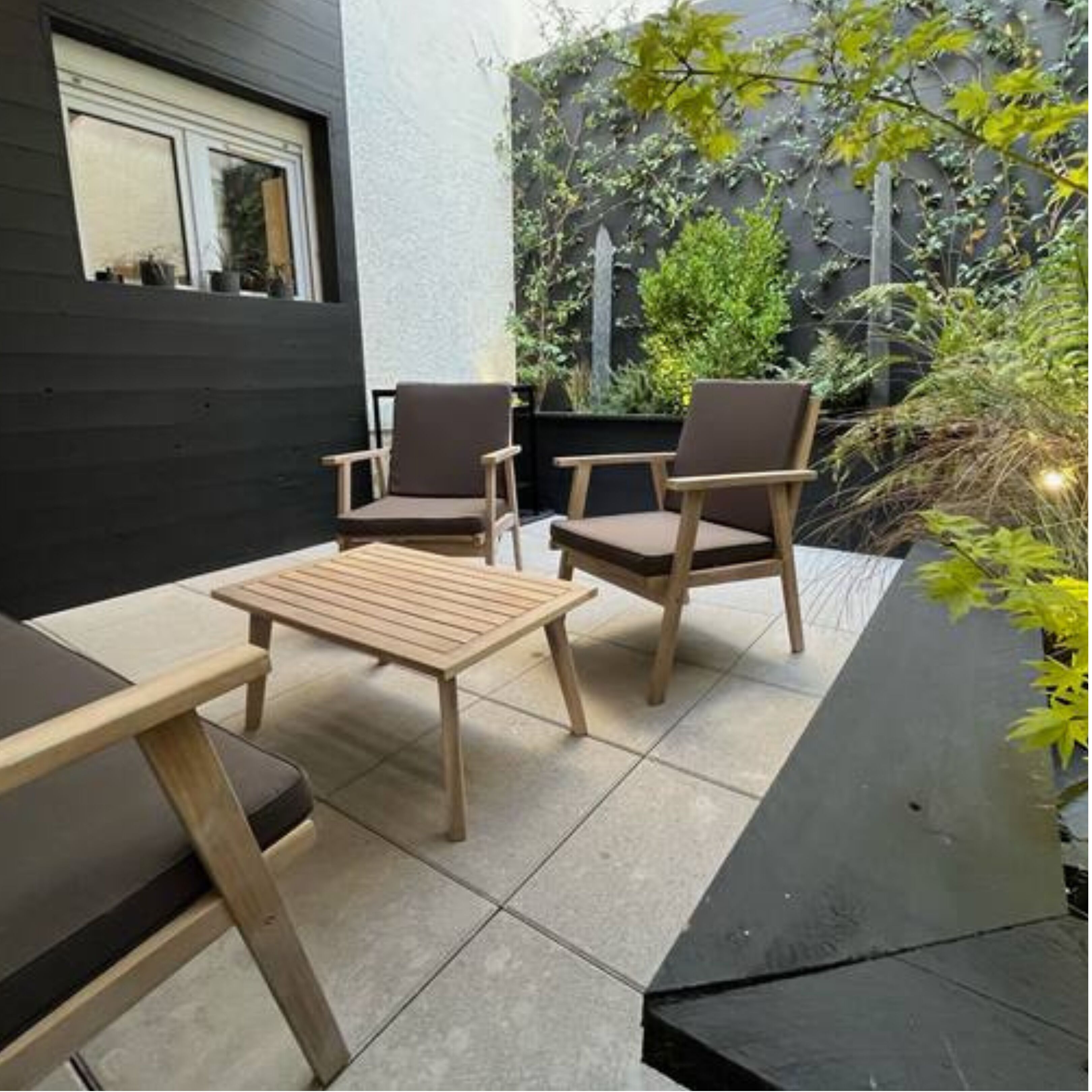 An outdoor seating area with two wooden chairs and a table, surrounded by a garden with various plants and a black wall.