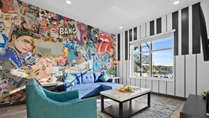 A modern living room with a vibrant comic-style mural, a blue sofa, teal armchairs, and a large window with a city view.