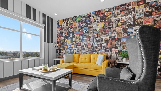 A modern living room with a yellow sofa, a gray armchair, and a coffee table. A large wall mural of various posters and images decorates the space.