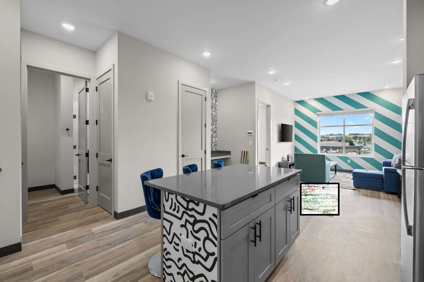 Una cocina moderna con una isla central, un comedor con sillas azules y una sala de estar con un ventanal grande.