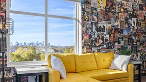 Una habitación con un sofá amarillo, un ventanal grande y una pared decorada con distintos carteles.