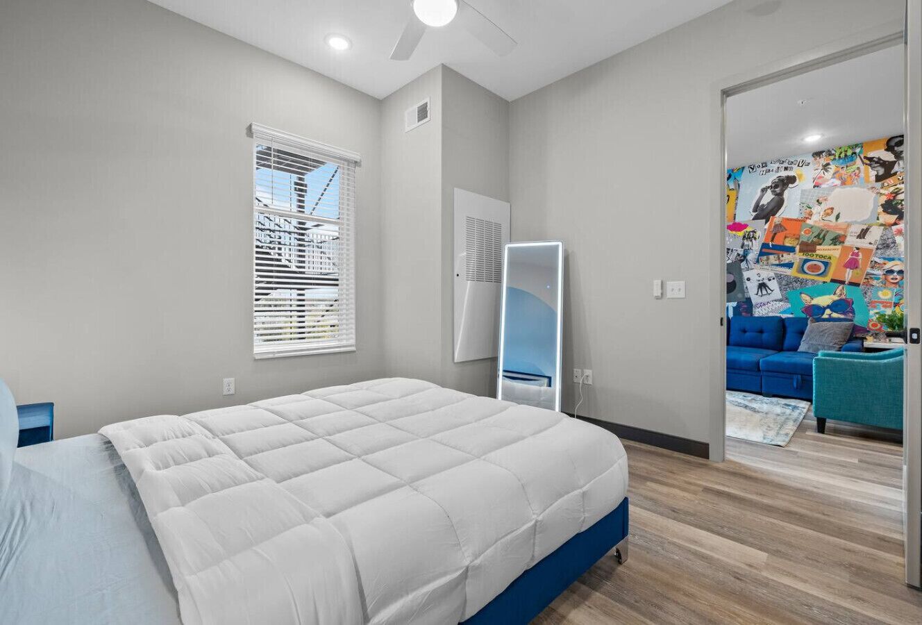 Un dormitorio con una cama grande, un ventilador de techo, una ventana con persianas, un espejo y un colorido adorno en la pared de la habitación contigua.