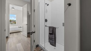 A modern bathroom with a glass shower enclosure, a towel rack, and a visible door leading to a bedroom with a bed and a window.