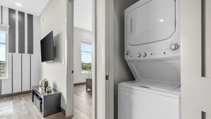 A modern laundry room with a front-loading washing machine, a flat-screen TV mounted on the wall, and a small table with a radio and decorative items. - 1BR Haven – Quick Drive to Honky Tonks & More! (Nashville)