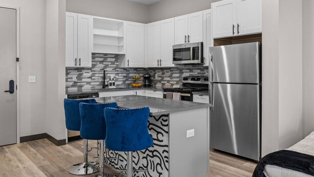 A modern kitchen with white cabinets, a stainless steel refrigerator, a microwave, and a gas stove. There are two blue bar stools at the island. - Cozy 1BR Escape – Close to the Heart of Nashville! (Nashville)