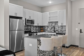 A modern kitchen with white cabinets, stainless steel appliances, a tiled backsplash, and a central island with bar stools. - Stay Close to the Action – Cozy 1BR Nash Retreat (Nashville)