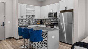 Una cocina moderna con armarios blancos, un refrigerador de acero inoxidable, un microondas y una estufa a gas. Hay dos taburetes azules en la isla.