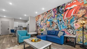 Un salón moderno con un mural vibrante de estilo cómic, un sofá azul, una mesa de centro de mármol y una cocina al fondo.