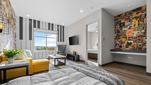 A modern bedroom with a large bed, a sofa, a coffee table, and a TV. There is a window with a view of the sky and a wall with a collage of images.