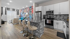 A modern kitchen with white cabinets, a central island with bar stools, a colorful mural, and a dining area with a blue sofa.