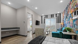 A modern hotel room with a large bed, a seating area with a sofa and armchair, a small table, and a colorful mural on the wall.