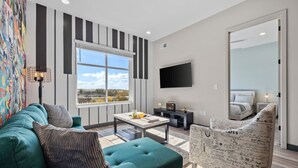 Un salon moderne avec un canapé, une table basse et un fauteuil à motifs. Un téléviseur à écran plat est fixé au mur, et une fenêtre offre une vue sur la ville.