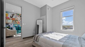Une chambre avec un lit, une fenêtre donnant sur un beau paysage, un miroir et une porte qui mène à un espace de vie comprenant un canapé et une fresque murale colorée.