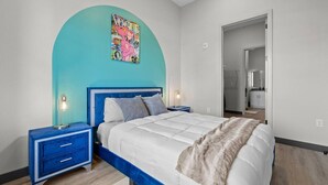 A bedroom with a bed, nightstands, a blue headboard, a wall art piece, and a doorway leading to a bathroom.