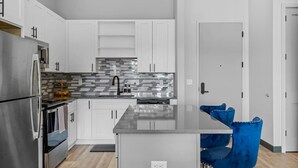 A modern kitchen with white cabinets, a stainless steel refrigerator, a tiled backsplash, and a central island with blue chairs.