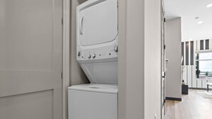 A modern laundry room with a front-loading washer and dryer, a door leading to a hallway, and a view of the outside through a window. - Urban Oasis 1BR - Quick Access to Downtown Nash! (Nashville)